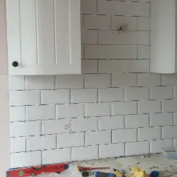 Układanie białych, prostokątnych płytek na ścianie w kuchni, widoczne krzyżyki dystansowe, poziomica, narzędzia glazurnicze na blacie i szafka kuchenna.