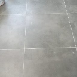 Wykończenia Przemysław Robert - Ułożone szare płytki podłogowe w łazience, widoczna fuga, narzędzia glazurnicze i resztki materiałów budowlanych w rogu pomieszczenia.