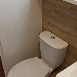 Kompaktowa toaleta w łazience wykończonej białymi i drewnopodobnymi płytkami, widoczne elementy instalacji sanitarnej i fragment umywalki.