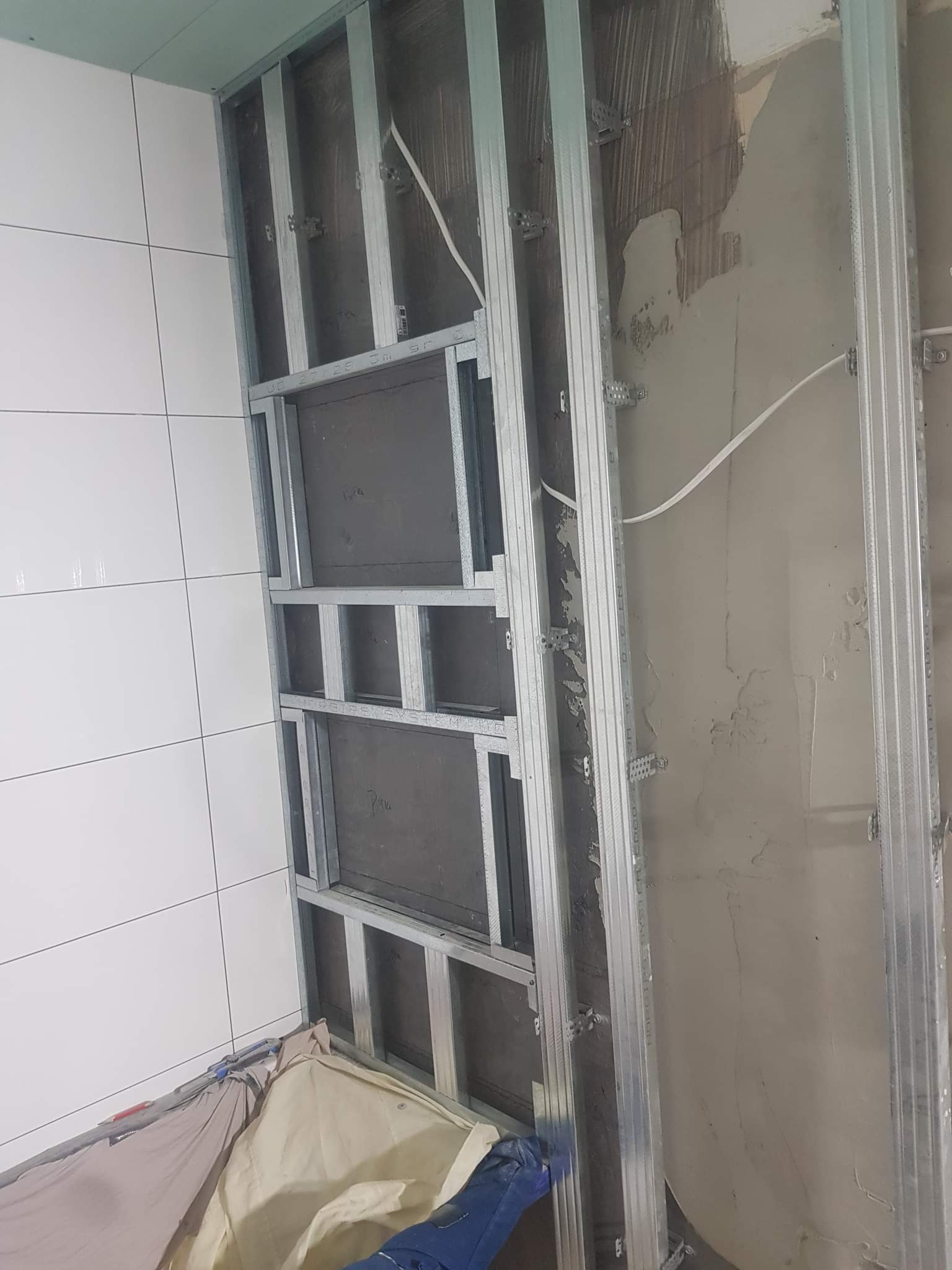 Montaż metalowego stelaża pod zabudowę w łazience, widoczne profile pionowe i poziome, przygotowane wnęki, fragment ściany z białymi płytkami oraz surowy fragment ściany z widocznymi śladami zaprawy.