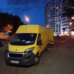 Żółty bus marki Peugeot z otwartymi drzwiami bocznymi, zaparkowany na chodniku z kostki brukowej przed wysokim budynkiem mieszkalnym o zmierzchu.