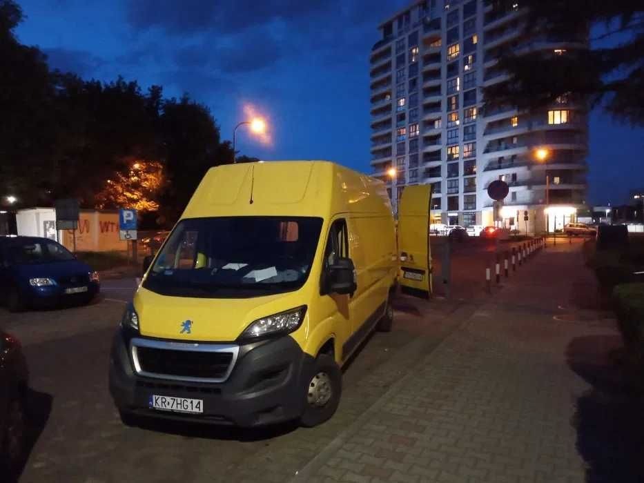 Żółty bus marki Peugeot z otwartymi drzwiami bocznymi, zaparkowany na chodniku z kostki brukowej przed wysokim budynkiem mieszkalnym o zmierzchu.