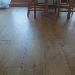 Ułożone panele podłogowe w odcieniach brązu, widoczne fragmenty nóg drewnianych krzeseł i zasłony w tle.