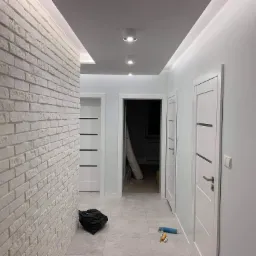 Korytarz po remoncie z białą cegłą na ścianie, trzema białymi drzwiami, szarą podłogą, workiem na śmieci i wałkiem malarskim na podłodze.