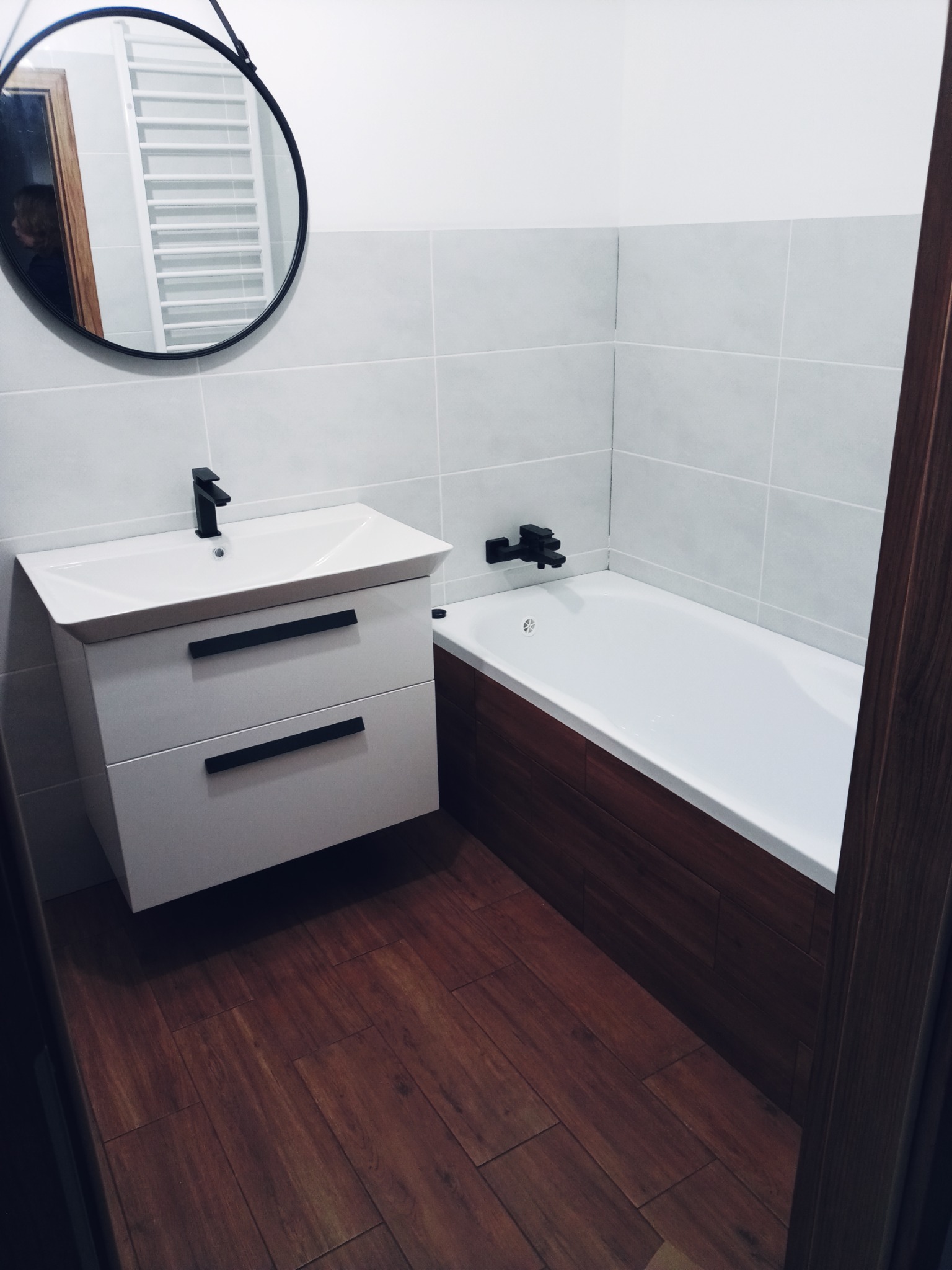 Nowoczesna łazienka z drewnianą podłogą i akcentami w kolorze czarnym: okrągłe lustro, bateria umywalkowa, uchwyty szafek i bateria wannowa.