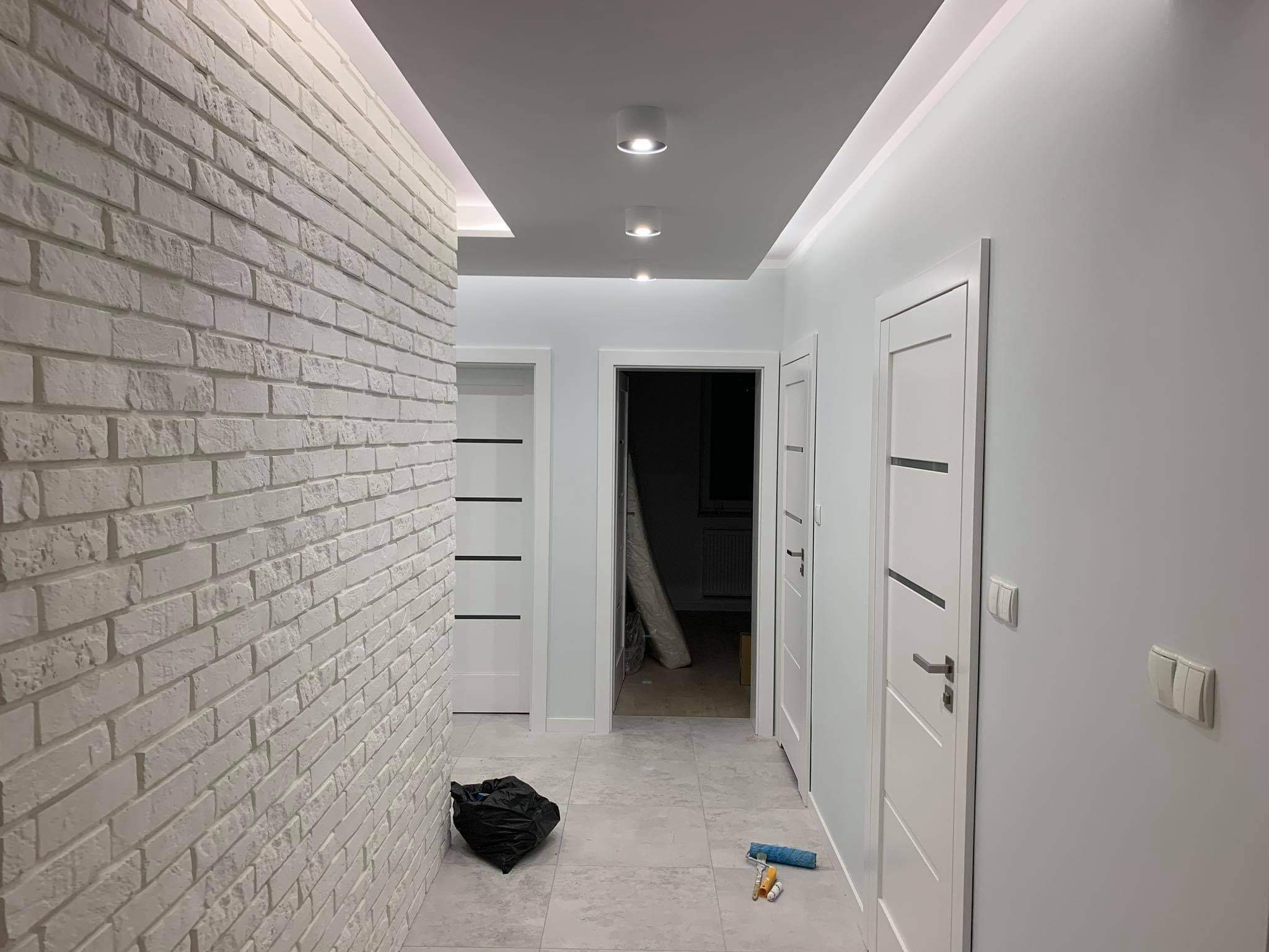 Korytarz po remoncie z białą cegłą na ścianie, trzema białymi drzwiami, szarą podłogą, workiem na śmieci i wałkiem malarskim na podłodze.