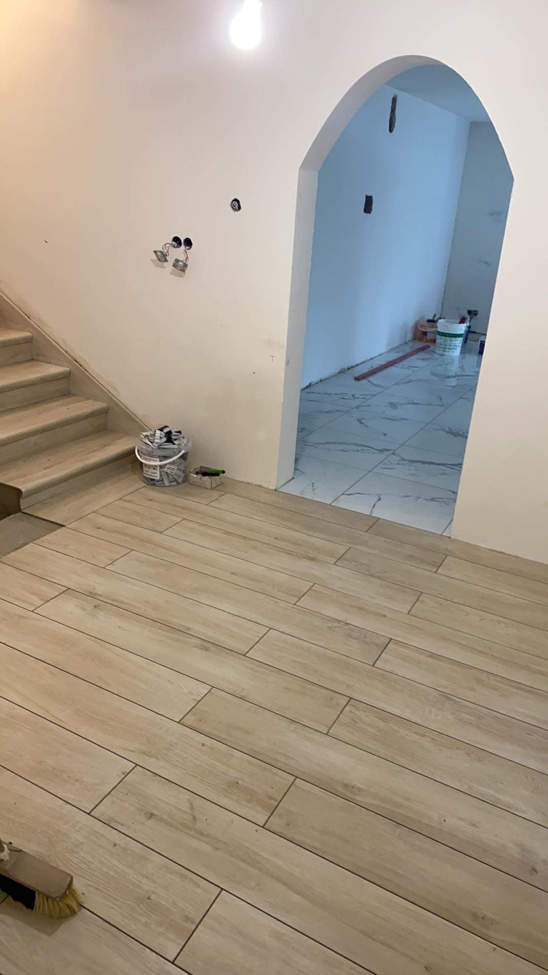 Ułożona podłoga z paneli imitujących drewno, widoczne schody z marmurowymi stopniami, przejście do pomieszczenia z marmurową podłogą, wiadro z narzędziami i szczotka w rogu.