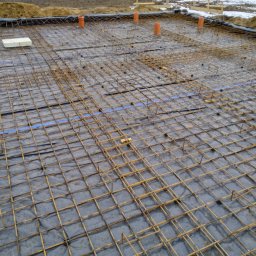 Deco-Bud Katarzyna Biernat - Przygotowanie zbrojenia fundamentu: siatka z prętów stalowych ułożona na folii hydroizolacyjnej, widoczne pionowe rury kanalizacyjne i fragment terenu z resztkami śniegu w tle.