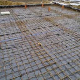 Przygotowanie zbrojenia fundamentu: siatka z prętów stalowych ułożona na folii hydroizolacyjnej, widoczne pionowe rury kanalizacyjne i fragment terenu z resztkami śniegu w tle.