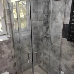 Szklana kabina prysznicowa z deszczownicą i panelami z szarych płytek imitujących kamień, widok z zewnątrz na fragment okna z zielenią.