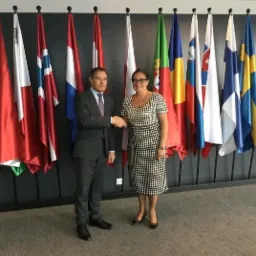 Uścisk dłoni pomiędzy mężczyzną w garniturze a kobietą w sukience na tle flag państwowych, symbolizujący zawarcie umowy lub partnerstwo.