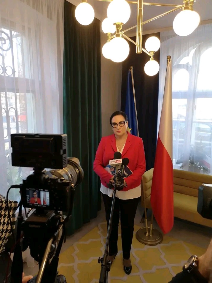 Kobieta w czerwonej marynarce udziela wywiadu przed kamerą, z mikrofonami kilku stacji telewizyjnych, na tle flagi Polski i Unii Europejskiej.