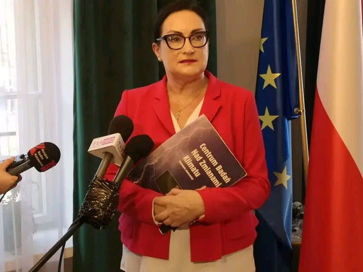 Kobieta w czerwonej marynarce trzyma książkę 'Centrum Badań Nad Zmianami Klimatu' przed mikrofonami stacji radiowych i telewizyjnych, z flagami UE i Polski w tle.