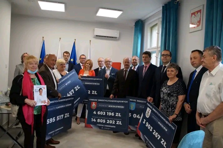 Grupa osób prezentuje symboliczne czeki z kwotami dotacji dla różnych gmin i powiatu Mikołowskiego, wręczane w jasnym pomieszczeniu z flagami UE i Polski w tle.