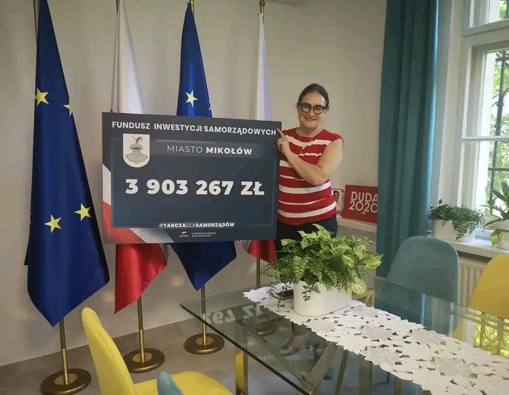 Kobieta w okularach prezentuje tablicę z informacją o dofinansowaniu dla Miasta Mikołów w kwocie 3 903 267 zł z Funduszu Inwestycji Samorządowych, na tle flag państwowych i UE.