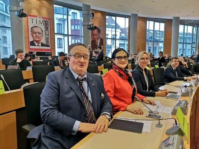 Sala konferencyjna z uczestnikami siedzącymi przy stołach, w tle plakaty z portretami