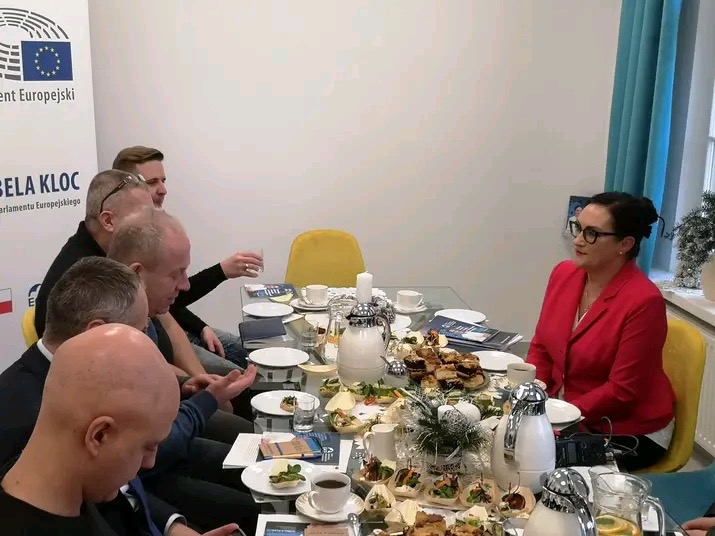 Grupa osób w trakcie spotkania biznesowego przy stole z przekąskami i napojami, w tle flaga Unii Europejskiej