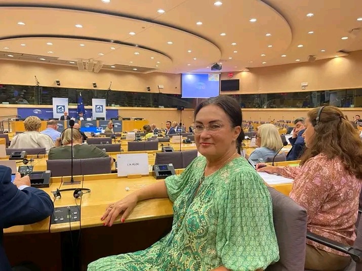 Kobieta w zielonej sukience siedzi przy stole z mikrofonem w sali konferencyjnej z innymi osobami w tle.