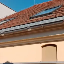 Budynek z pomarańczową dachówką, dwoma oknami dachowymi i metalową rynną z uchwytami, widok z dołu na tle błękitnego nieba.