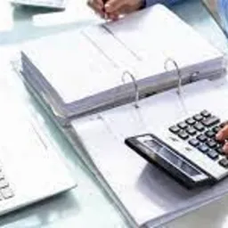 Osoba w błękitnej koszuli używa kalkulatora nad otwartym segregatorze z dokumentami; obok laptop, okulary i roślina w doniczce na biurku.