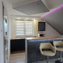 Nowoczesna kuchnia w aneksie z barem wykończonym imitacją kamienia, oświetleniem LED i dwoma krzesłami barowymi na tle okna z żaluzjami poziomymi.