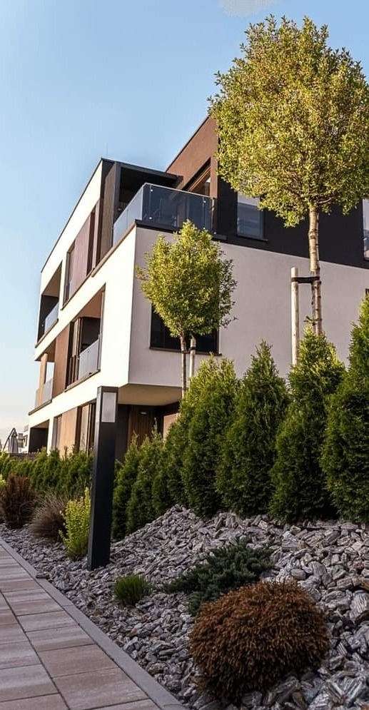 Nowoczesny budynek wielorodzinny z zagospodarowanym terenem: równo ułożona kostka brukowa, ozdobne kamienie, tuje i drzewa podparte palikami.