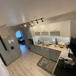 Szare szafki kuchenne z drewnianym blatem i białymi płytkami na ścianie, widok na salon przez łukowate przejście