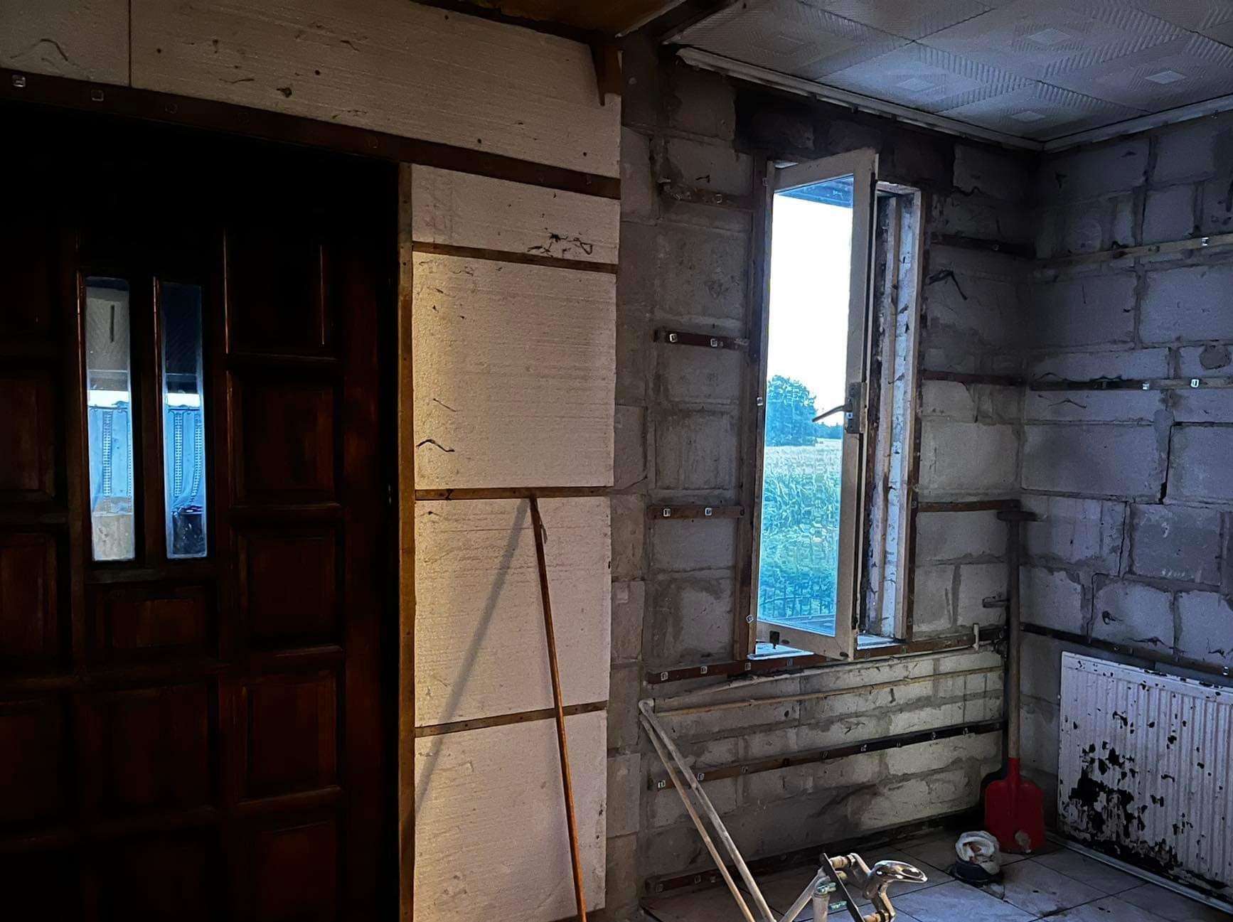 Wnętrze w trakcie remontu: ściany z surowego betonu komórkowego, częściowo ocieplone styropianem, okno z widokiem na pole, stara drewniana stolarka drzwiowa, grzejnik z odpryskami farby, widoczne...
