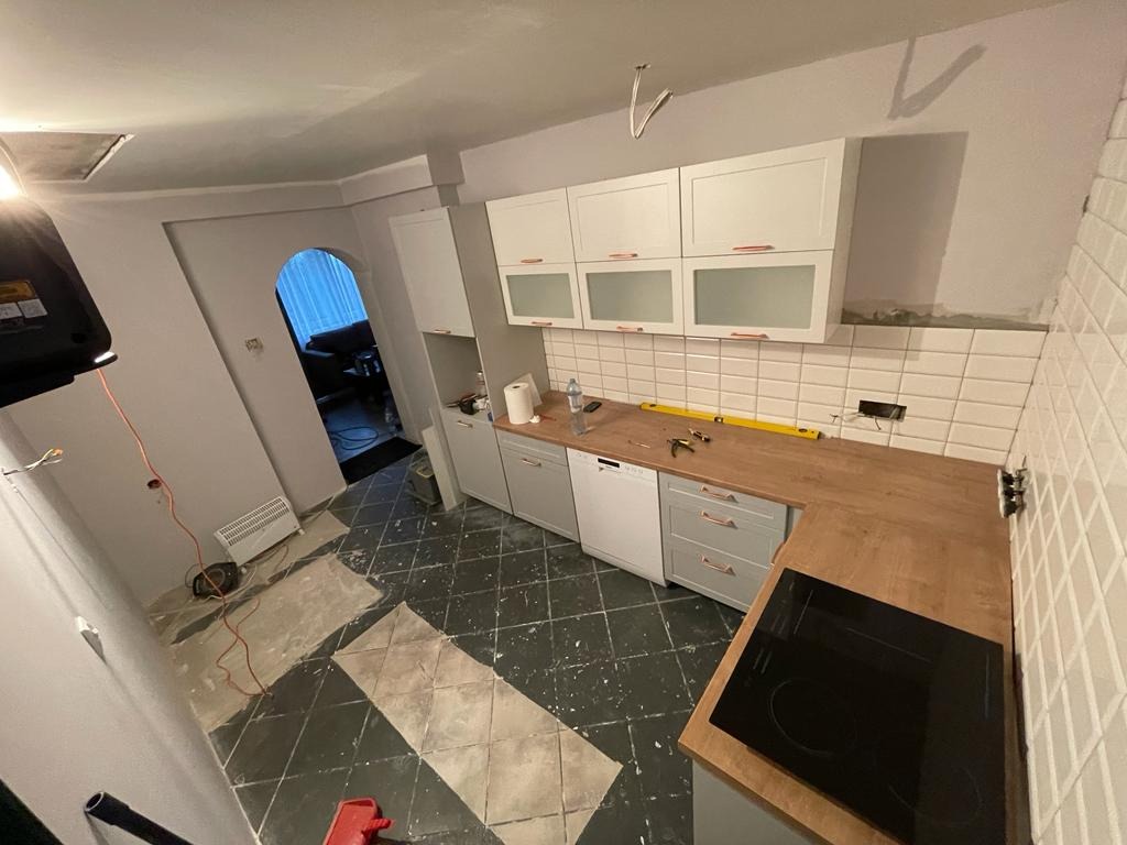 Wnętrze kuchni w trakcie remontu: białe szafki wiszące, drewniany blat roboczy, częściowo ułożone płytki na ścianie, czarna płyta indukcyjna, podłoga z czarno-białą szachownicą, widoczne narzędzia...