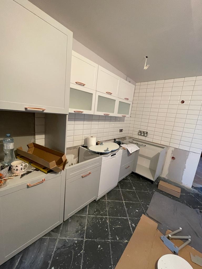Montaż szarych szafek kuchennych z miedzianymi uchwytami, widoczne białe płytki na ścianie, kartony i akcesoria montażowe na podłodze.
