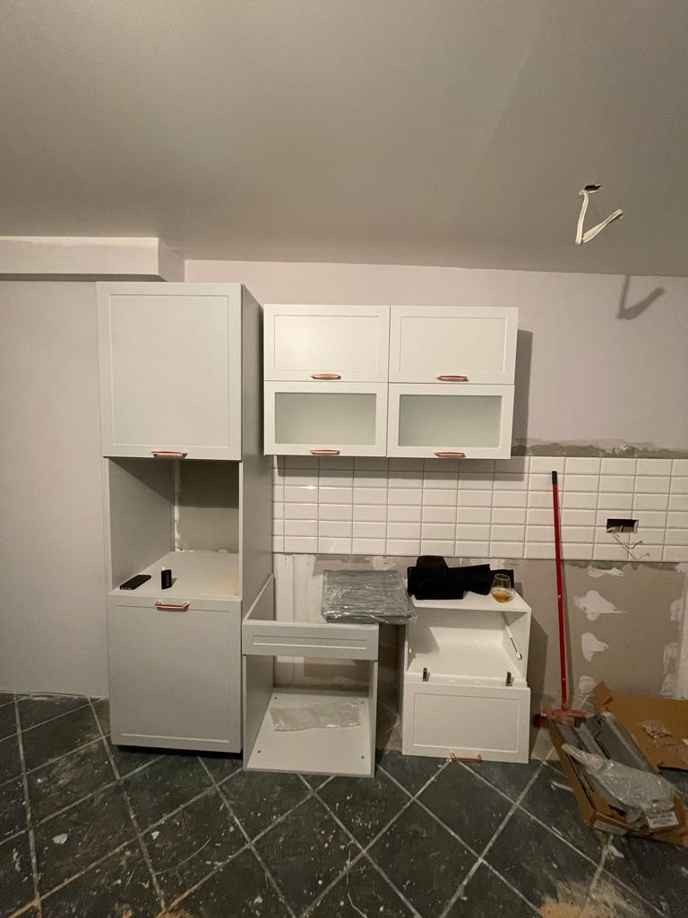 Montaż białych szafek kuchennych z miedzianymi uchwytami na tle białych płytek i surowej ściany, widoczne elementy instalacji elektrycznej i narzędzia.