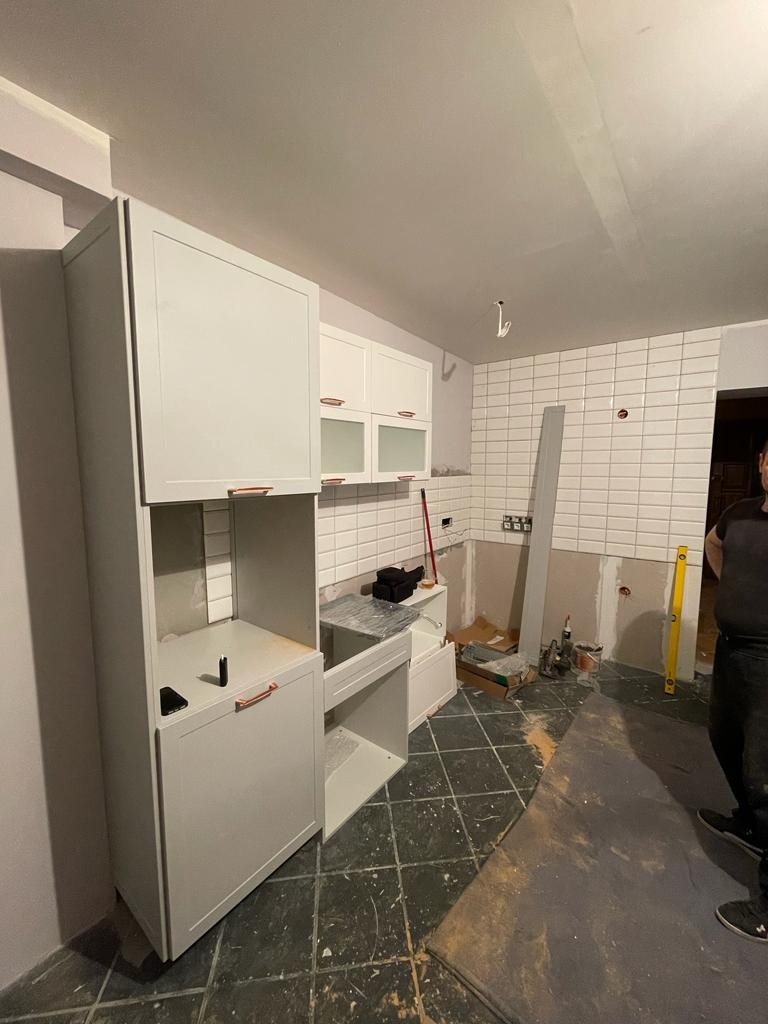 Montaż białych szafek kuchennych z miedzianymi uchwytami w trakcie remontu kuchni, widoczne płytki na ścianie, poziomica i narzędzia.