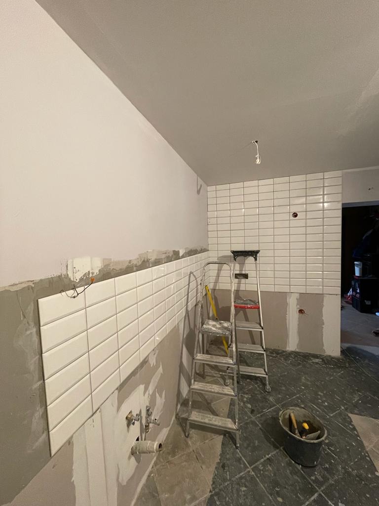 Ściana w trakcie wykańczania białymi, prostokątnymi płytkami, widoczna drabina i wiadro z narzędziami na podłodze z ciemnych płytek.