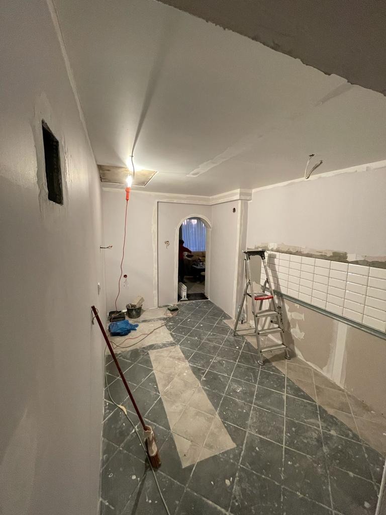 Wnętrze w trakcie malowania ścian na szaro, widoczny fragment ułożonych białych płytek, drabina, oraz czarno-biała podłoga w romby z fragmentami szarej wylewki.