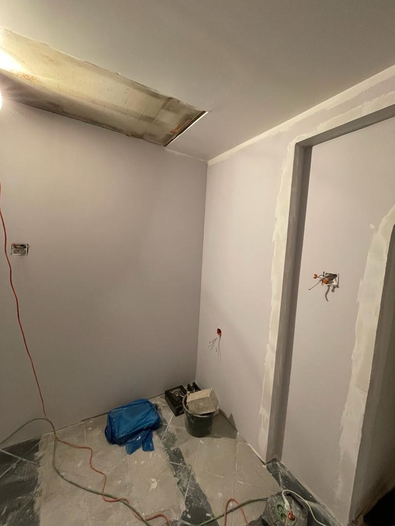Pokój w trakcie remontu: szare ściany po tynkowaniu, widoczne kable elektryczne, otwór w suficie, wiadro z zaprawą, niebieska folia ochronna na podłodze.