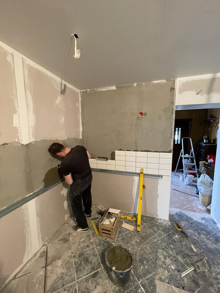 Mężczyzna w czarnej koszulce i spodniach układa białe, prostokątne płytki na ścianie w pomieszczeniu w trakcie remontu, widoczny poziomica, wiadro z zaprawą i częściowo ułożone płytki w rzędzie.