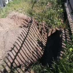 Wykopana dziura w ziemi obok trawy i ogrodzenia, oświetlona słońcem rzucającym cienie sztachet na ziemię.