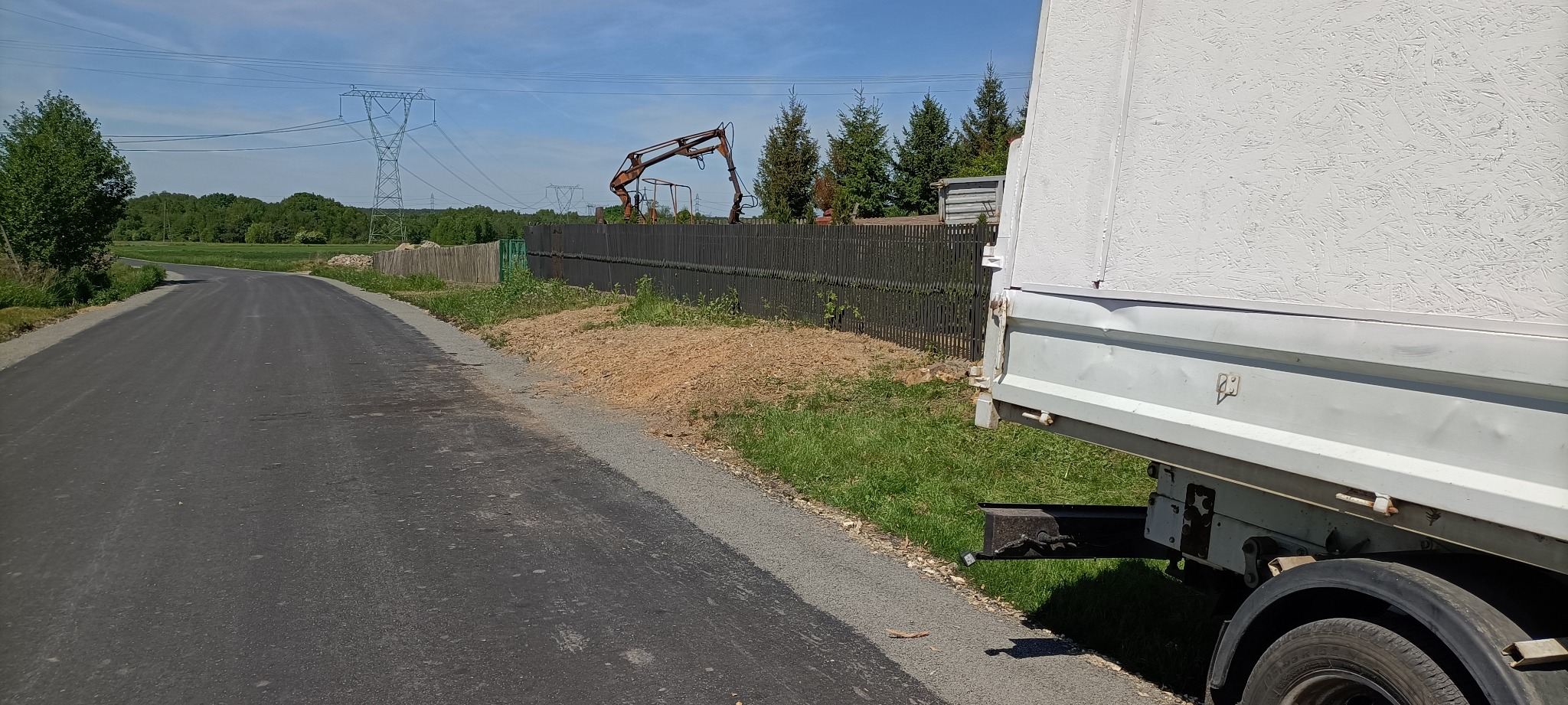 Ciężarówka zaparkowana przy asfalcie, w tle drewniany płot i wysięgnik koparki