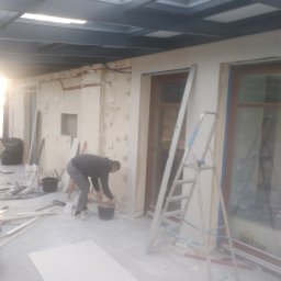 Michał Rzeczkowski - Prace przygotowawcze do wykończenia ścian w jasnym pomieszczeniu z oknami, widoczna drabina, płyta gipsowa i osoba nakładająca masę szpachlową na ścianę.