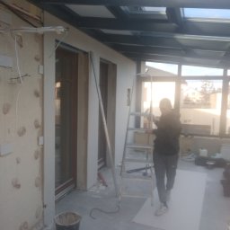 Michał Rzeczkowski - Prace wykończeniowe w zabudowie balkonowej z przeszklonym dachem, widoczna osoba na drabinie obok przygotowanej ściany z widocznymi przewodami elektrycznymi i wiaderkiem na podłodze.