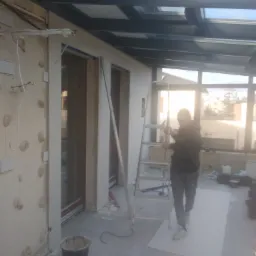 Prace wykończeniowe w zabudowie balkonowej z przeszklonym dachem, widoczna osoba na drabinie obok przygotowanej ściany z widocznymi przewodami elektrycznymi i wiaderkiem na podłodze.