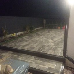 Michał Rzeczkowski - Ułożona, prostokątna kostka brukowa imitująca drewno na zewnątrz budynku, widoczne narzędzia i materiały przygotowane do dalszych prac.