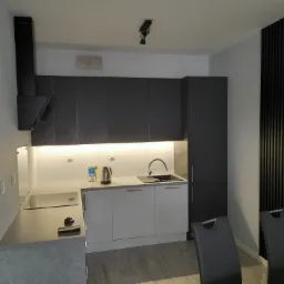 Nowoczesna kuchnia z szarymi szafkami górnymi i białymi dolnymi, stalowym czajnikiem i czarnym okapem, oświetlona ledowym pasem pod szafkami, z czarną ścianą dekoracyjną z pionowych listew i dwoma...