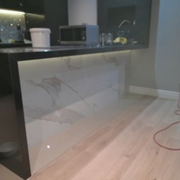 Michał Rzeczkowski - Nowoczesna zabudowa wyspy kuchennej z białymi płytami imitującymi marmur, podświetlona od dołu, z widocznym fragmentem drewnianej podłogi i sprzętem do czyszczenia podłóg.