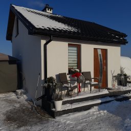 Michał Rzeczkowski - Biały dom z czarnym dachem pokrytym śniegiem, widoczne trzy krzesła na schodach, dekoracje świąteczne i kot pod krzesłem, zimowy krajobraz