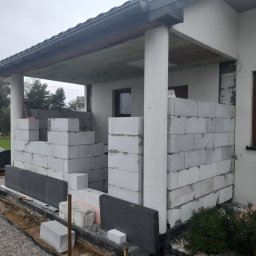 Michał Rzeczkowski - Wznoszenie ścian z bloczków betonowych na zadaszonym tarasie domu jednorodzinnego, widoczne fragmenty izolacji pionowej i poziomej, narzędzia pomiarowe oraz przygotowane bloczki.