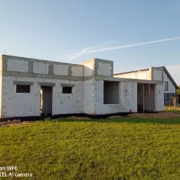 Surowy stan domu jednorodzinnego z betonu komórkowego, widoczne otwory okienne i drzwiowe, fundament zabezpieczony czarną folią, w tle sąsiednie domy i zielony trawnik.