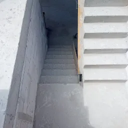 Surowe betonowe schody prowadzące do piwnicy w trakcie budowy, widoczne szalunki i ślady po deskach na ścianach.