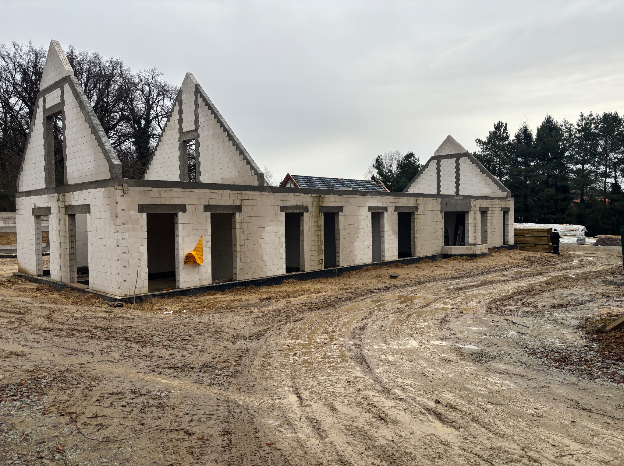 Surowy stan domu z białych bloczków, widoczne otwory okienne i drzwiowe, teren budowy błotnisty po deszczu, w tle drzewa i fragment dachu sąsiedniego budynku.