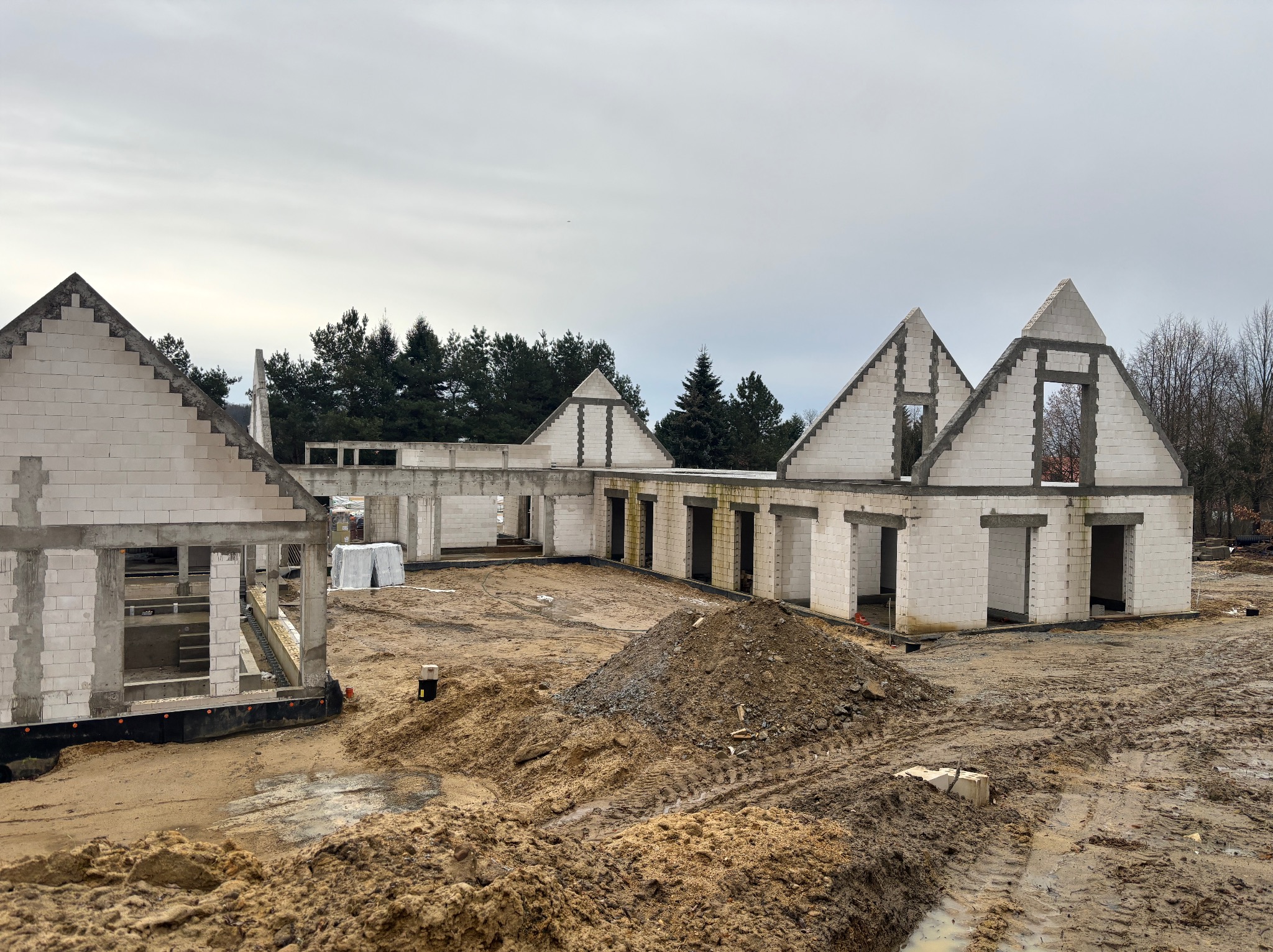 Surowy stan budowy domu z białych bloczków, widoczne ściany i otwory okienne, na pierwszym planie hałda ziemi i błoto.